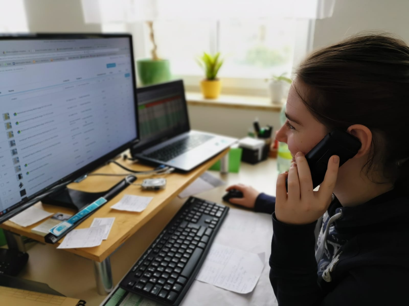 Eine Person mit langen Haaren und einem dunklen Kapuzenpullover sitzt an einem Schreibtisch, schaut auf einen Computermonitor voller Daten und spricht mit einem Smartphone. Am Arbeitsplatz befinden sich eine Tastatur, eine Maus, Haftnotizen und eine Pflanze am Fenster.