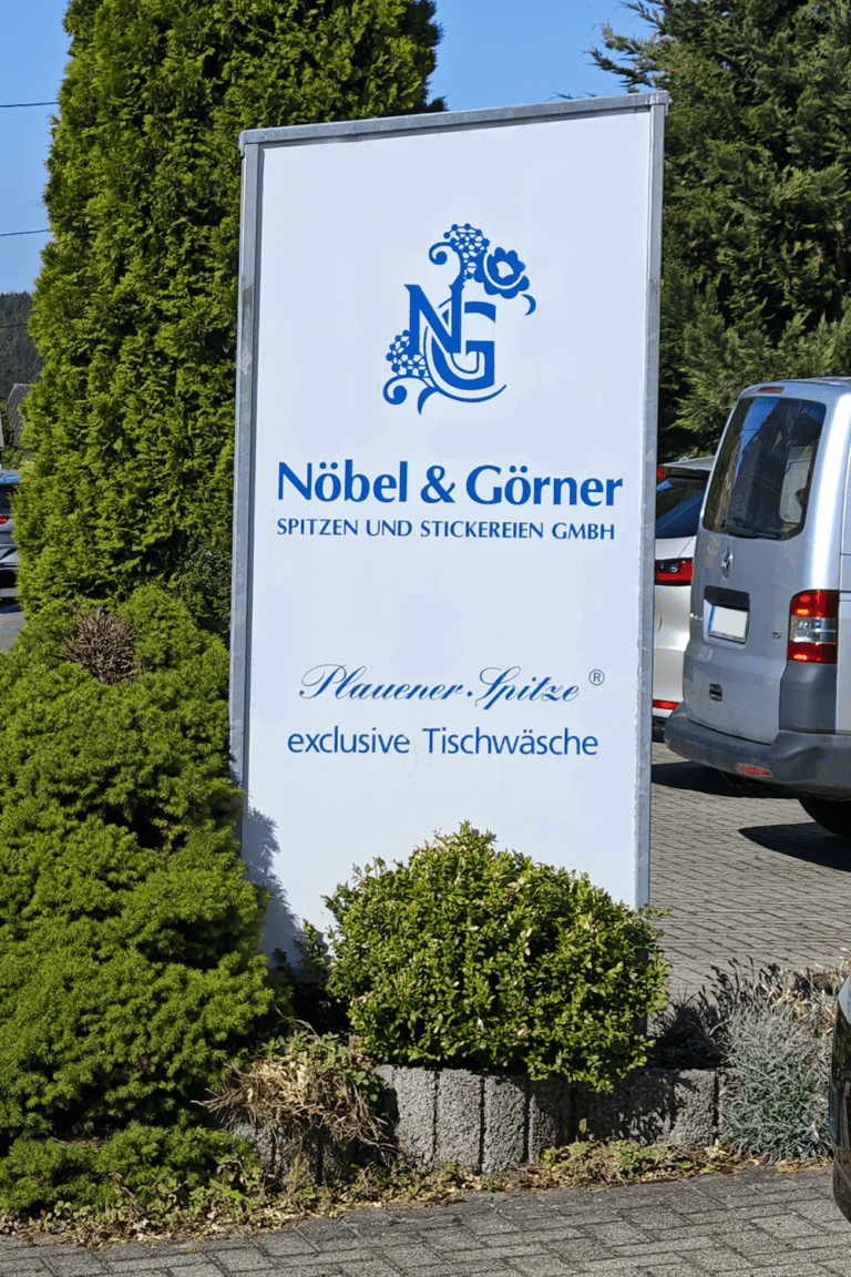 Schild der „Nöbel & Görner Spitzen und Stickereien GmbH“ mit Logo und Schriftzug „Plauener Spitze exklusive Tischwäsche“ neben geparkten Fahrzeugen und Grünflächen.