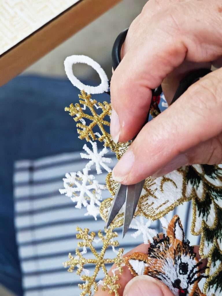 Ein Paar Hände schneidet sorgfältig mit einer Schere komplizierte, gestickte Ornamente aus. Die Ornamente umfassen goldene und weiße Schneeflocken und ein kleines Design mit einem Fuchs. Im Hintergrund ist teilweise ein blau-weiß gestreiftes Tuch sichtbar.