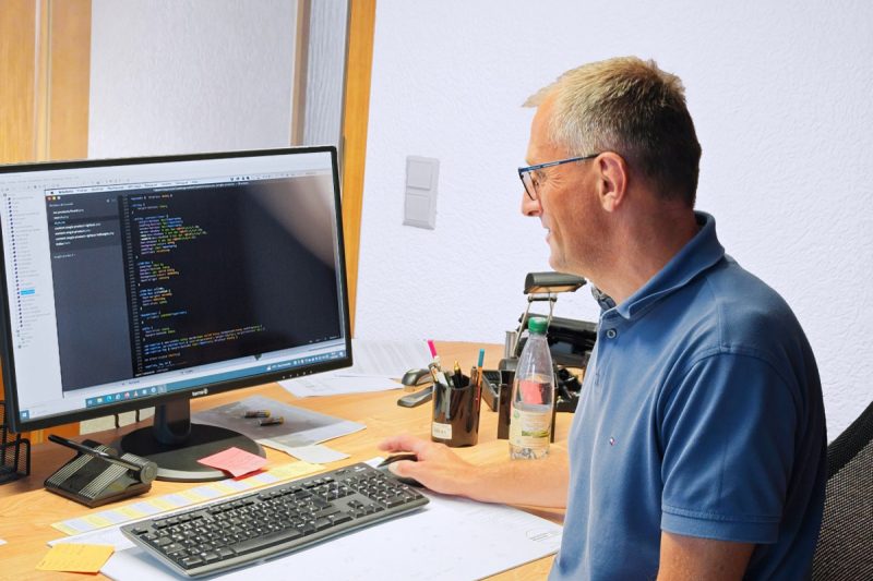 Jens Nöbel in einem blauen Poloshirt sitzt an einem Schreibtisch und arbeitet an einem Computer. Auf dem Bildschirm sind bunte Codezeilen zu sehen. Auf dem Schreibtisch liegen eine Tastatur, Papiere, Haftnotizen, Stifte und eine Wasserflasche herum.