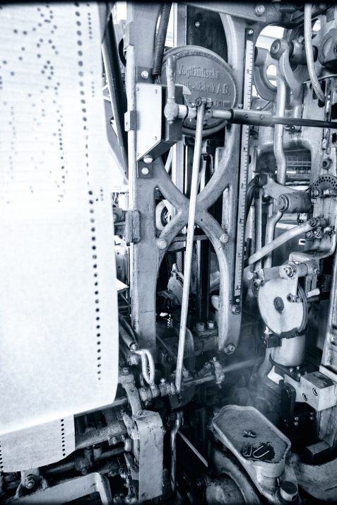 Eine Schwarz-Weiß-Nahaufnahme einer komplexen, historischen mechanischen Maschine mit Zahnrädern, Hebeln und Stangen. Links ist eine Rolle perforierten Papiers zu sehen, was darauf hindeutet, dass die Maschine Daten oder Anweisungen verarbeitet. Die dichte und komplizierte Mechanik erinnert an die Präzision der Geräte von „Nöbel und Görner“, die zur Herstellung der Plauener Spitze verwendet werden – einer feinen Spitzenarbeit, die für ihre aufwendigen Spitzen und Stickereien bekannt ist.